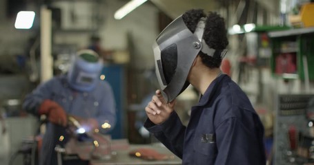 4K Young apprentice welder lifting visor & smiling at camera in workshop. Slow motion