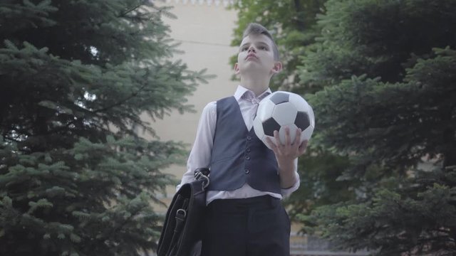 Handsome Well-dressed Boy Standing On The Street Holding The Soccer Ball And Purse. Serious Young Man Simultaneously Acting Like Child And Adult.