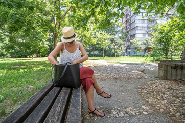 Young fashionable girl with hat sitting on the bench in the park trying to find something phone or...