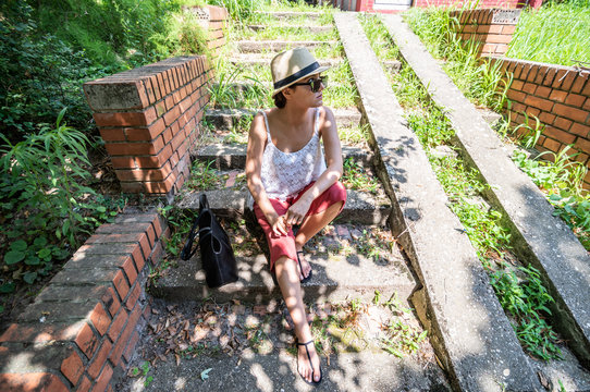 Young hipster girl with hat in retro or vintage look clothes sitting on the stairs in the park in shade on hot sunny summer day for rest and taking a break