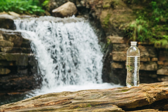 Transparent plastic bottle with water stands on a wooden trunk against the background of the river and the waterfall. The concept of pure natural drinking water