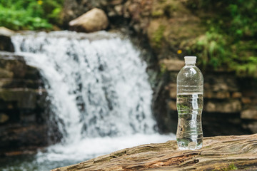 Transparent plastic bottle with water stands on a wooden trunk against the background of the river and the waterfall. The concept of pure natural drinking water