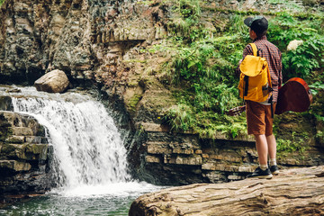 Traveler man with a backpack and a guitar stands on a trunk of a tree against a waterfall. Space for your text message or promotional content