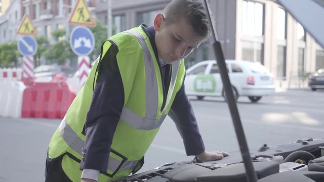 Confident little boy wearing safety equipment standing by the open hood of a broken car. Boy repairing a car. Child as adult.