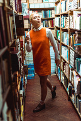 Obraz na płótnie Canvas beautiful and blonde woman in glasses and orange dress looking away in library