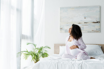 happy young pregnant african american woman sitting on bed and touching belly