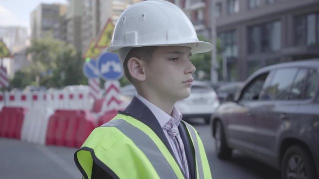 Portrait cute little boy wearing business suit and safety equipment and constructor helmet standing on a busy road in a big city. Engineer, architect, builder doing his work. Child as adult.