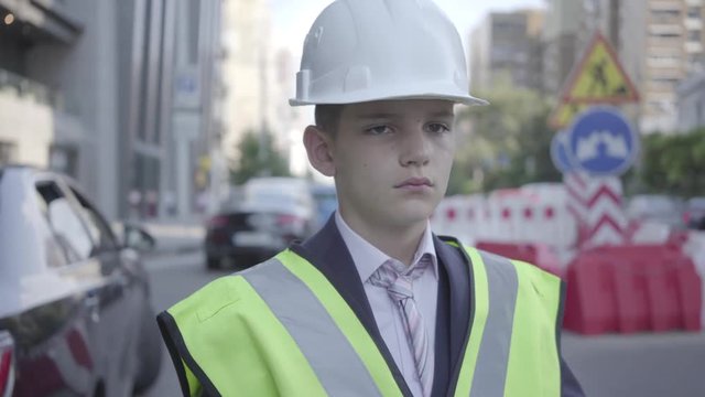 Portrait cute little boy wearing business suit and safety equipment and constructor helmet standing on a busy road in a big city. Engineer, architect, builder doing his work. Child as adult.