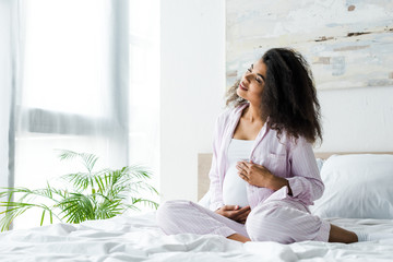 cheerful young pregnant african american woman with closed eyes sitting on bed and touching belly