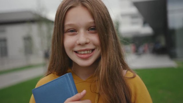 Portrait of a cheerful girl student enjoying her time at school. Close-up of a teenager smiling broadly holding a book staying outdoors during a break near campus.
