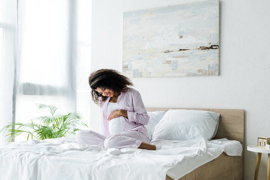 attractive pregnant african american woman looking at belly and sitting on bed