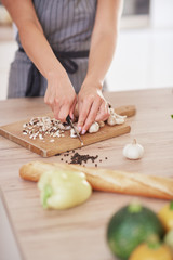 Dedicated Caucasian housewife in apron standing in kitchen and chopping mushrooms. On table are lots of vegetables. Cooking at home concept.