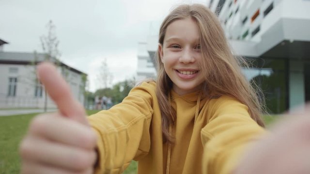 Portrait of a pretty cute girl taking selfie outdoors near the campus. Close-up of positive happy teenage student smiling and showing thumb up on camera.