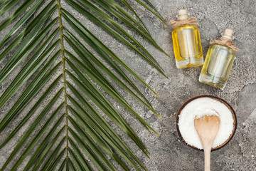 top view of coconut shavings with wooden spoon near oil in bottles on grey textured background with palm leaf