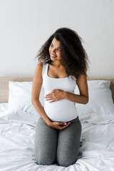 positive curly african american pregnant woman touching belly while sitting on bed