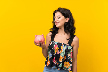 Young woman over isolated yellow background holding a big piggybank