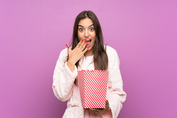 Young woman in pajamas and dressing gown over isolated purple background holding popcorns