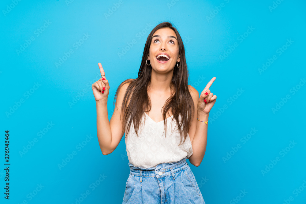 Wall mural Young woman over isolated blue background surprised and pointing up