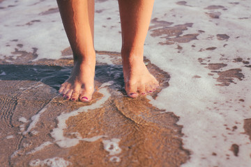 Obraz na płótnie Canvas Female legs in the water. The girl goes along the coastline. Sunset light and sea foam.