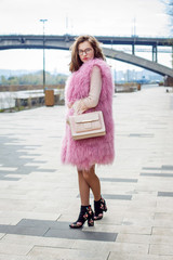 Young woman in a pink fur vest with a light bag, posing in the spring outside and smiling.