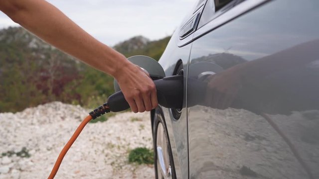 Woman is charging an electric car at mountains background and fresh air