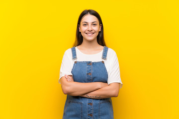 Young woman in dungarees over isolated yellow background keeping the arms crossed in frontal...
