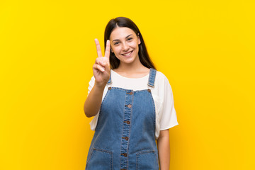 Young woman in dungarees over isolated yellow background smiling and showing victory sign