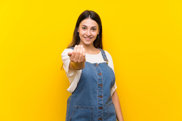 Young woman in dungarees over isolated yellow background inviting to come with hand. Happy that you came