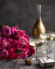 Two glasses of champagne with a bottle behind, a bouquet of purple peonies, small present box