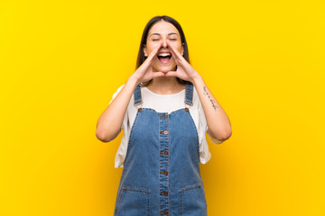 Young woman in dungarees over isolated yellow background shouting and announcing something