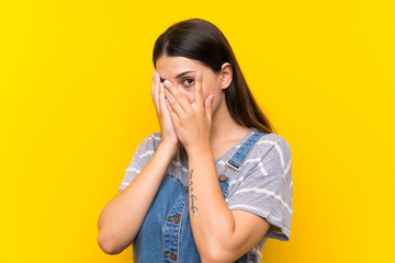 Young woman in dungarees over isolated yellow background covering eyes and looking through fingers