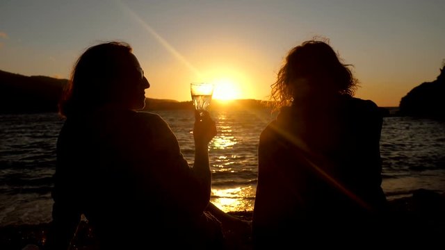 Silhouette of two women sitting on shore of Mediterranean sea, drinking an alcoholic drink at sunset. Greece. Slow motion. HD