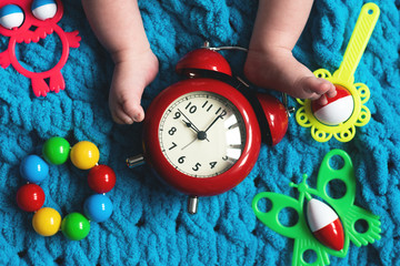 Baby feet and a red alarm clock and child toys on a blue towel background.