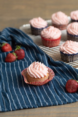 Homemade strawberry cupcakes in red and blue paper cups on cooling rack.  One isolated in front on blue paper towel.  Fresh strawberries scattered around.