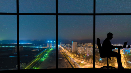 The man working at the table near a panoramic window with a night city view
