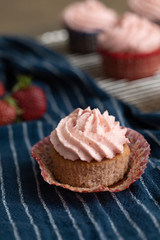 Homemade strawberry cupcakes in red and blue paper cups on cooling rack.  One isolated in front on blue paper towel.  Fresh strawberries scattered around.