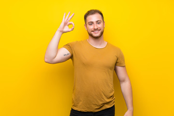 Fototapeta premium Blonde man over isolated yellow wall showing ok sign with fingers