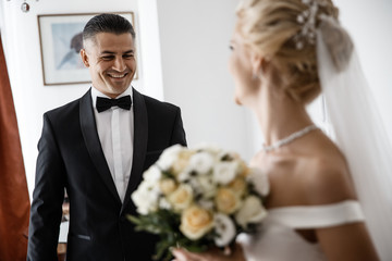 A groom and a bride are looking at each other. A bride is holding a bouquet and a groom is laughing.