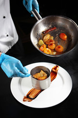 The cook lays out the cooked peach flambe from the pan on a plate. There is a special form on the plate. Concept food show in a restaurant. Photos in the interior.
