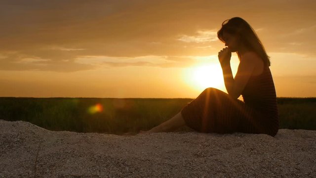 silhouette of a young woman on the sand at sunset, girl bowed her head in prayer to the Creator with gratitude, the concept of rest, religion, the beauty of nature and harmony