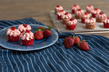 Homemade mini strawberry cupcakes in red and white paper liners.  Some isolated on blue plate with fresh strawberries.  Some cooling on rack in background.  Blue kitchen towel on wood table.