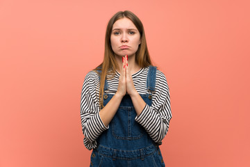 Young woman with overalls over pink wall keeps palm together. Person asks for something