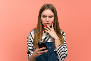 Young woman with overalls over pink wall with a mobile and thinking