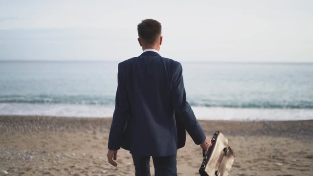 A man in a business suit is walking along the beach towards the sea