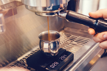 Closeup of professional cappuccino espresso machine making coffee. Thin hot drink liquid stream flow coming pouring to metal mug jug standing on scale. Toned with warm yellow film filters.