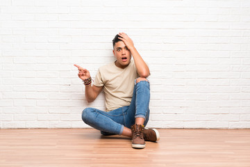 Young man sitting on the floor surprised and pointing finger to the side
