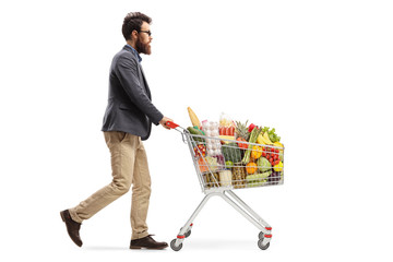 Bearded guy walking and pushing a shopping cart with food products - obrazy, fototapety, plakaty