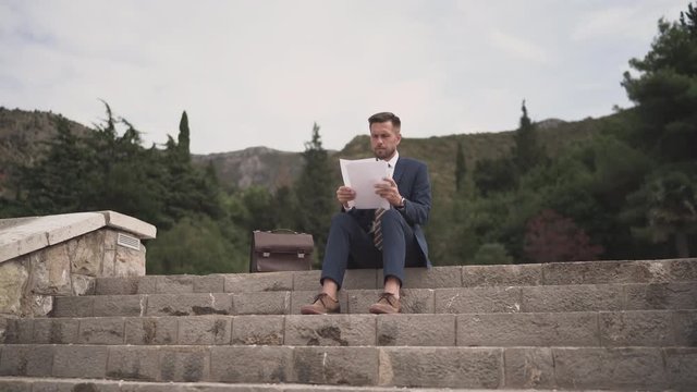 Businessman sits on the top step and scrolls through contracts, reads documents. 4K