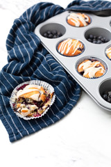 Freshly-baked lemon-glazed blueberry muffins in a muffin tin.  One isolated in front broken open.  Blue Kitchen towel on white countertop.