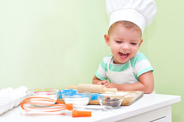 Cheerful cute baby boy in a chef costume laughs standing at the table where doing cookies.	